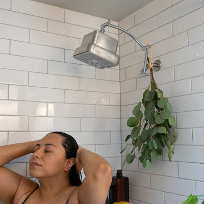 Woman in a shower with white tiled walls and a plant hanging from the shower head extender which is raising the showerhead up higher