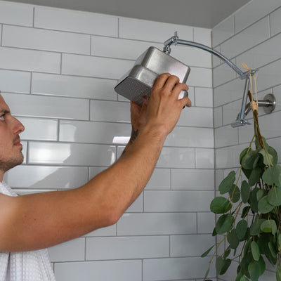 Man in a shower with white tiled walls and a plant hanging from the shower head extender which is raising the showerhead up higher